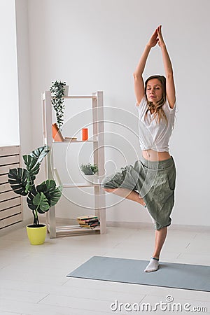 Woman practise yoga performs. Pose on mat inside of cozy room with plants and greenery. Healthy lifestyle concept. Stock Photo