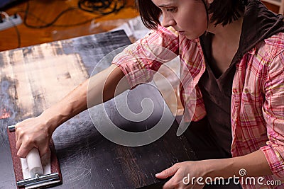 Woman prepares the surface of an old wooden black table for painting, using a carpentry holder for sandpaper. Home Stock Photo