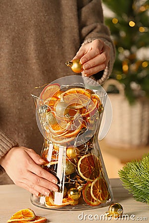 Woman putting Christmas tree ball into glass vase with dry orange slices and cones at white wooden table, closeup, Festive decor Stock Photo