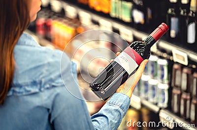 Woman reading the label of red wine bottle in liquor store or alcohol section of supermarket. Shelf full of alcoholic beverages. Stock Photo