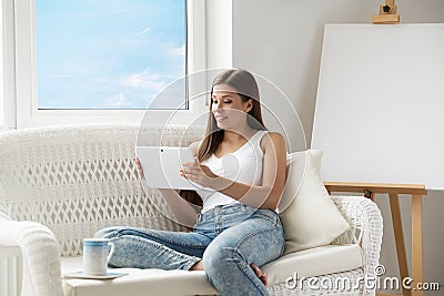 Woman reading Tablet PC sitting on Sofa in front of empty Easel Canvas. Young Girl using Touch Pad Screen resting next to Window. Stock Photo