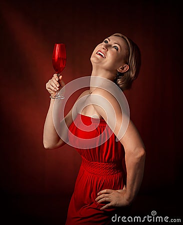 A woman in a red dress stands on a brown background and holds a wine glass in her hand. Middle aged blonde. Stock Photo