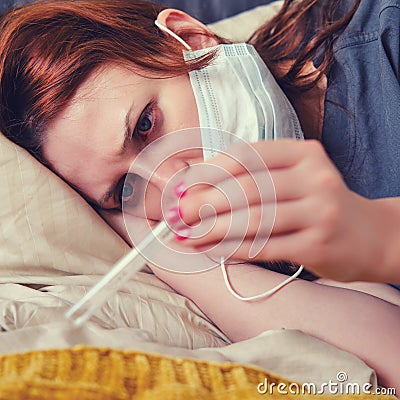 A woman with red eyes, sick with the flu, looks sadly at the thermometer. The patient lies on a bed in a medical mask Stock Photo