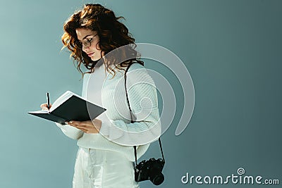 Woman with red hair writing in notebook while standing with digital camera on grey Stock Photo