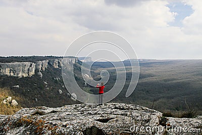 A woman in a red sweater stands on top of a mountain and photographs a mountain landscape. Stock Photo