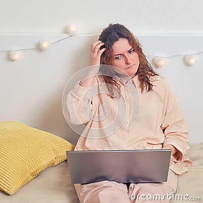 Woman reflected while sitting on the bed with a laptop. Sad girl with question on her face while working online from home Stock Photo
