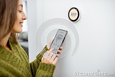 Woman regulating heating temperature with phone and thermostat at home Stock Photo