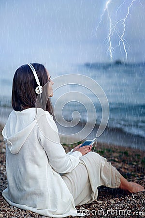 Woman relax listening music under rain, sitting on a sea beach. Stock Photo