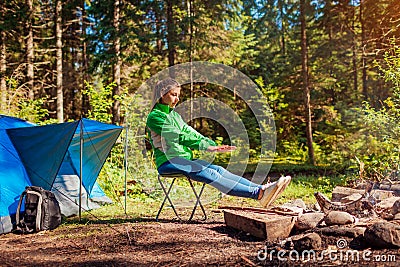 Woman relaxing by campfire in forest sitting next to tent warming hands and feet. Summer camping. Traveling alone Stock Photo