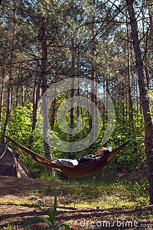 Woman relaxing in the hammock in the middle of a pine forest, watching sundown. Slow life concept. Hipster. Camping tent. Vertical Stock Photo