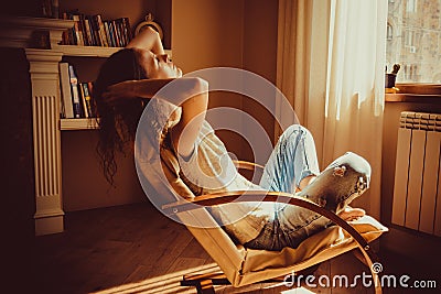 Woman relaxing after work in comfortable modern chair near window in livingroom. Warm natural light. Cozy home. Casual clothing. C Stock Photo
