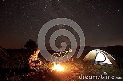 Woman resting at night camping near campfire, tourist tent, bicycle under evening sky full of stars Stock Photo