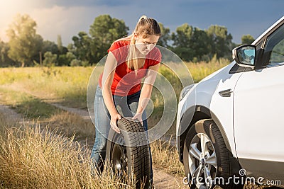 Young woman rolling spare wheel to change flate tyre on her car Stock Photo