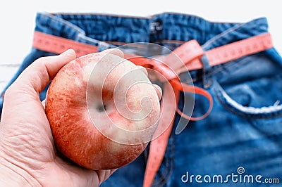 A woman`s hand holds an Apple against the top of a pair of denim trousers on a wooden background. Blue jeans with a measuring tap Stock Photo