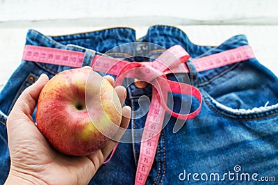 A woman`s hand holds an Apple against the top of a pair of denim trousers on a wooden background. Blue jeans with a measuring tap Stock Photo