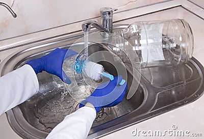 Womanâ€™s hands with gloves washing dish with fresh water at kitchen faucet over the metal sink. Human washes glass jar. Close up Stock Photo