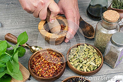 Woman`s hands grind red pepper in a wooden mortar. Kitchen gray table with various spices Stock Photo