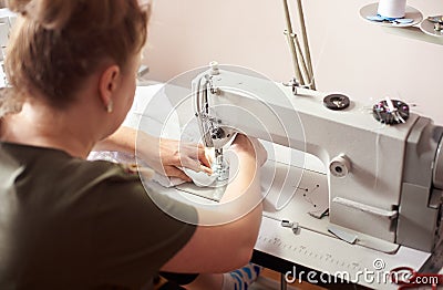 Woman seamstress sitting on workplace in atelier doing stitching white dress with professional sewing machine. Back view Stock Photo