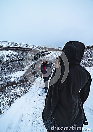 Woman shelters from the extreme cold, taking a photo of her husband in a totally snowy and mountainous environment in Iceland with Editorial Stock Photo