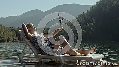 Woman shooting on handheld film gimbal stabilization for smartphone. Girl lie sunbed on pier make selfie. Blogger Stock Photo