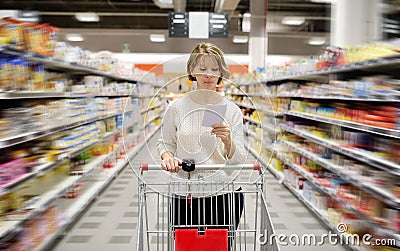 Woman with shopping list pushing cart looking at goods in supermarket Stock Photo