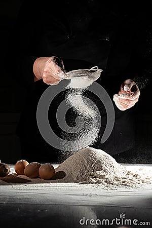 woman sifts flour through sieve onto table with eggs on black background. Stock Photo