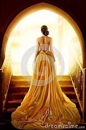 Woman Silhouette in Golden Luxury Gown. Elegant Lady in Yellow Long Silk Dress with naked Back Rear Side View looking at Light Stock Photo