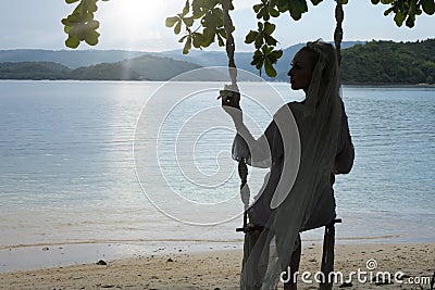Woman silhouette in a veil of bride and white short dress on a rope swing on background of the sunrise sea and islands on the ho Stock Photo