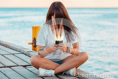 A woman sits on a pier by the sea and watches social media on her smartphone. Face smudged, sucked into the phone. The concept of Stock Photo