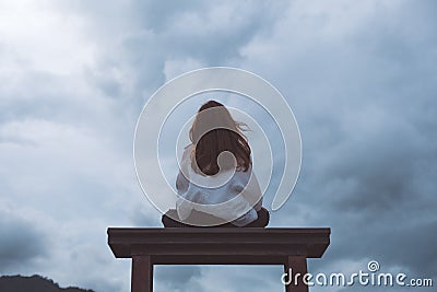 A woman sitting alone on a wooden bench in the park with cloudy and gloomy sky Stock Photo