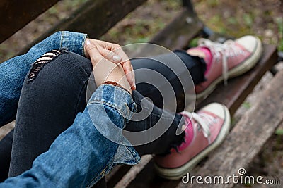 Woman sitting on a bench, holding her leg with her arms, with black ripped jeans and pink canvas sneakers Stock Photo