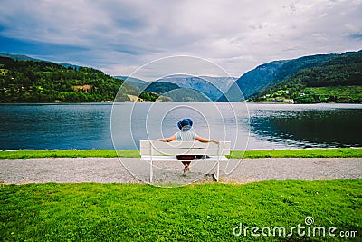 Woman sitting on a bench looking at the fjord in Ulvik, Norway. Fjord coastal promenade in Ulvik, Hordaland county, Norge. Lonely Stock Photo