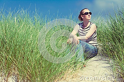 Woman sitting in sand dunes amongst tall grass relaxing, enjoying the view on sunny day Stock Photo