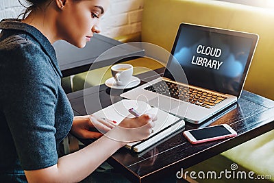 Woman sitting at table in cafe in front of laptop computer with inscription on monitor-cloud library and making notes in notebook Stock Photo