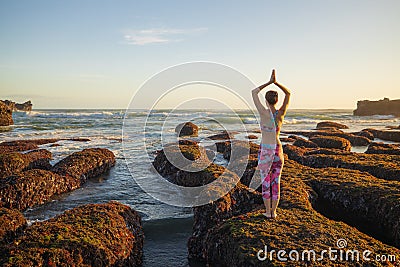 Woman standing on big stone during low tide, practicing yoga. Young woman raising arms with namaste mudra at the beach. Bali. View Stock Photo