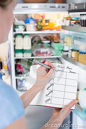 Woman Standing In Front Of Refrigerator In Kitchen With Notebook Writing Weekly Meal Plan Stock Photo