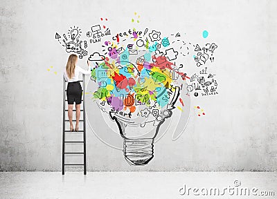 Woman standing on a ladder and drawing a large and colorful light bulb sketch on a concrete wall. Stock Photo