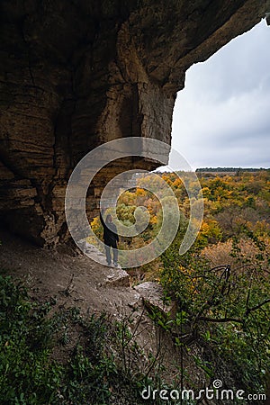 Woman standing by lighted exit in cave, daytime. Atmospheric snapshot in natural rock formations with autumn forest and dramatic Stock Photo