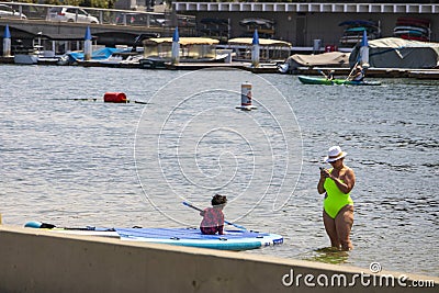 A woman standing in the ocean water using a cell phone with a child on a paddle board and people rowing kayaks in Long Beach Editorial Stock Photo