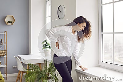 Woman with lumbago pain standing by the window sill and holding her hand on her lower back Stock Photo