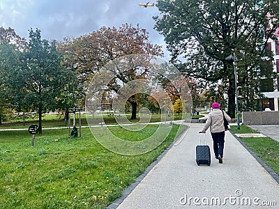 Woman with a suitcase is walking through the park. There's an airplane flying over her Stock Photo