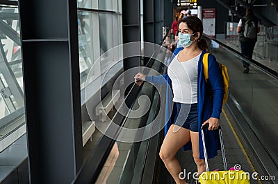 A woman with a suitcase is wearing a mask on the escalator at the airport. Stock Photo