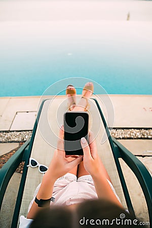 Woman sunbathing in chair by the pool and using mobile phone in hands Stock Photo