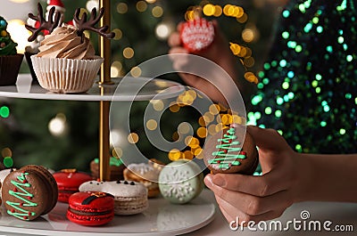 Woman taking Christmas macaron from stand against blurred festive lights, closeup Stock Photo