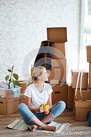 Woman with tea in hand sitting on floor of new apartment, pile of moving boxes on background Stock Photo