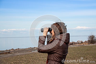 Woman tourist looking through binoculars at distant sea, enjoying landscape. Autumnal time.Lonely woman in brown coat Stock Photo