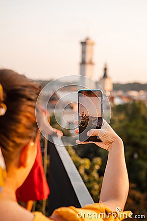 woman tourist looking at city on sunset from observation deck taking picture on her phone Stock Photo