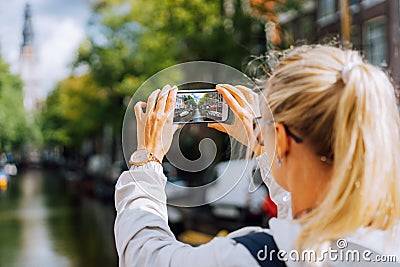 Woman tourist taking a picture of canal in Amsterdam on the mobile phone. Warm gold afternoon sunlight. Travel in Europe Stock Photo