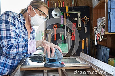 Mature Woman Upcycling Furniture In Workshop At Home Using Electric Sander Stock Photo