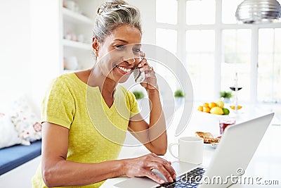 Woman Using Laptop And Talking On Phone In Kitchen At Home Stock Photo